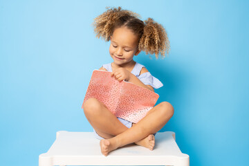 Cute little girl sitting on a table writing in notebook isolated over blue background. Back to...