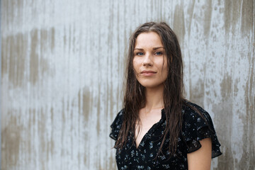 Fototapeta premium Simple portrait of a woman model next to the blank grey wall. Candid portraits of authentic people. Real life. Authentic people wearing black dress. Natural light portrait outdoors