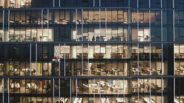 Cinematic drone shot at the glass offices of a skyscraper close-up at night, the camera flies back from the building. Aerial view of offices and business people working late.