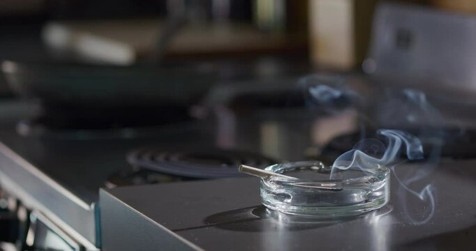 Interior detail shot of ashtray that has a lit marijuana CBD pre-roll joint, cigarette with smoke rising sitting in kitchen next to stove top