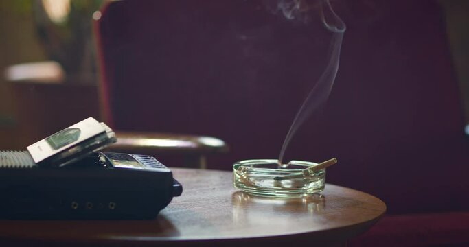 Low angle home interior detail shot of side table with vintage tape recorder / answering machine and ashtray that has a lit CBD marijuana pre-roll joint, cigarette with smoke rising