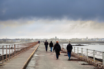 People walking on a Mutton causeway, Galway city in the background. Ireland. Selective focus. Outdoor activity. Cloudy sky. Town buildings in the background and dome of Galway Cathedral.