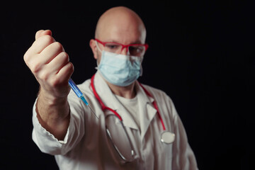 Portrait of a bald doctor with syringe full with blue color vaccine. Dark background. Selective focus. Health care and vaccination concept. Man in white coat, wearing mask and stethoscope.