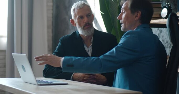 Two mature businessmen discussing business growth in modern office while sitting at desk. Man ceo talking with colleague about deal while meeting indoors