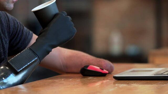 A caucasian man with amputated two stump hands is drinking coffee holding a cup with bionic hand and using the laptop with another limb. A man living with a disability is using the laptop