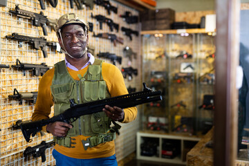African-american man wearing armor and helmet while standing with assault rifle in airsoft shop.