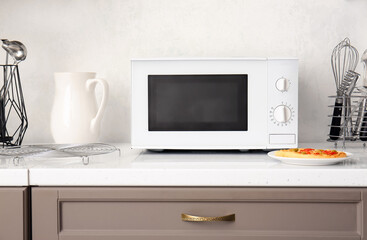 Modern microwave oven and plate with pizza on counter near light wall - obrazy, fototapety, plakaty