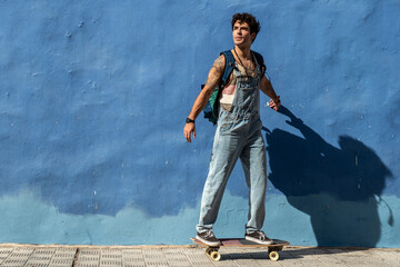 Stylish spanish guy hipster skateboarding on waveboard. City lifestyle. Blue wall.