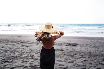 Fototapeta na wymiar Back view of beautiful woman with straw summer hat enjoying ocean breeze at the beach. Relax time.