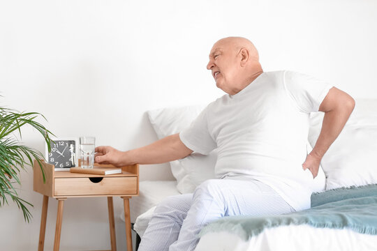 Senior man with back pain taking glass of water from table in bedroom
