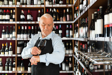 Confident cheerful elderly male winemaker holding glass of red wine, checking it in wine store