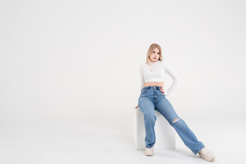 Young blonde girl in a white sweater and jeans sits and poses on an isolated white background in the studio. People lifestyle concept. Copy space for copy