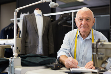 Elderly man tailor uses notepad to record jacket measurements in workshop