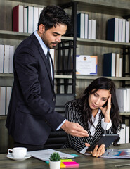 Millennial Indian Asian male successful executive businessman manager in formal suit holding clipboard discussing with female secretary businesswoman colleague at working desk in company office