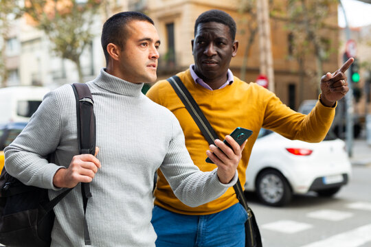 Caucasian man standing outdoors with smartphone and asking for directions African-american man.