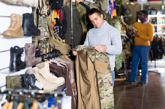 European man choosing camouflage clothes in military store. African-american man shopping in background.