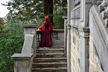 Fototapeta na wymiar portrait of pretty female model with red hair wearing glamorous renaissance red ballgown. Posing in a fairytale castle location with staircases 