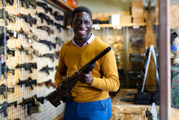 Positive african-american man standing in salesroom of arms shop with hunting rifle in hands and smiling.