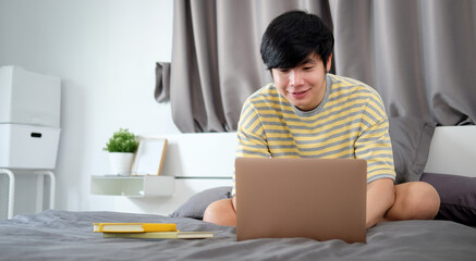 Handsome asian man sitting on bed and watching something on laptop computer.