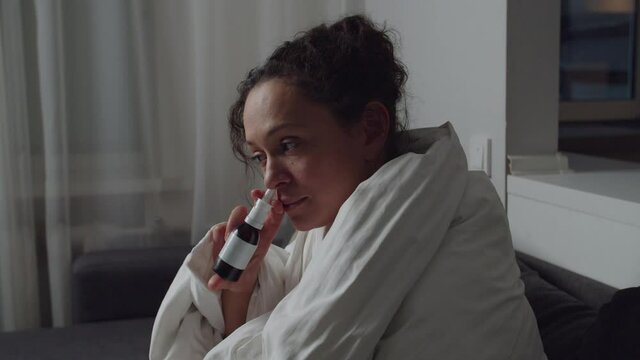 Face profile of sick adult female having runny nose, using nasal spray, wiping nose with paper napkin while sitting on couch indoors. Woman applying medicament drug curing disease at home. Moving shot