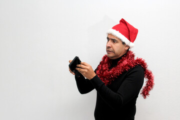 Latino adult man with hat and Christmas garland shows his wallet with very little money worried about not being able to celebrate Christmas
