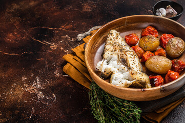 Baked halibut fish with roasted tomato and potato in wooden plate. Dark background. Top view. Copy...