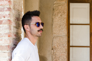 Fototapeta premium Young man in white shirt and sunglasses, standing leaning on the wall of a house