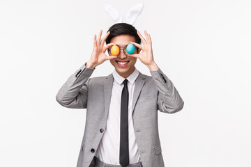 Holiday, people and celebration concept. Waist-up portrait of funny, playful and enthusiastic asian guy in grey suit and tie, holding painted eggs over eyes and smiling, enjoy Easter party