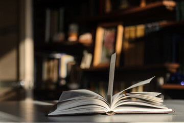 There is an open book on the table.  The background is bookshelves illuminated by the sun.