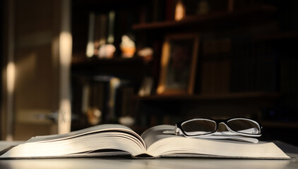 There is an open book on the table. There are glasses on the pages. The background is bookshelves illuminated by the sun.