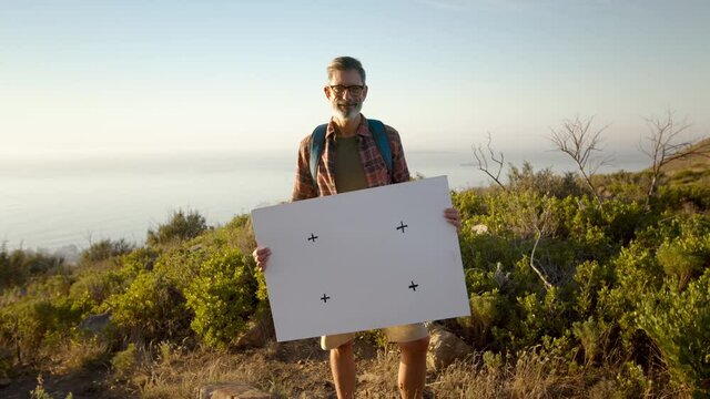 Mature hiker holding a blank placard on a hilltop. Happy mature backpacker displaying a banner while standing on top of a hill at sunset. Adventurous male activist smiling at the camera.