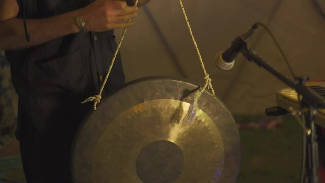 Close-up shot of a man wearing black shirt standing and playing with the gong in the lights background.
