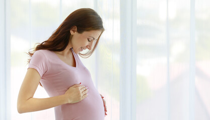 Caucasian young healthy female big belly tummy prenatal pregnant mother model in casual shirt standing in front of curtain background smiling holding glass of drinking milk in morning alone at home