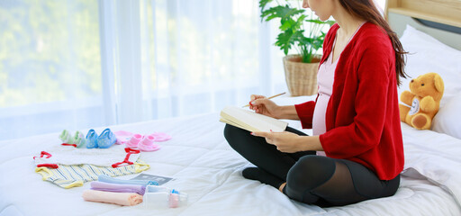 Caucasian young happy healthy female prenatal pregnant mother model in casual outfit with jacket sitting on bed in bedroom writing checklist note in notebook when preparing baby outfit and equipment