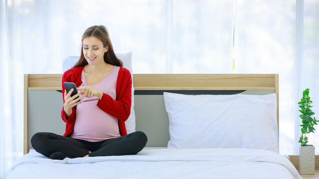 Caucasian millennial young happy healthy female prenatal pregnant mother in casual outfit sitting smiling on bed in bedroom via video call with smartphone.