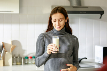 Caucasian millennial young happy female prenatal pregnant mother in casual outfit standing alone holding hand on big belly tummy with love drinking fresh milk for breakfast in full equipment kitchen