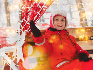 Christmas happy child portrait with bokeh lights festive decorations. Little girl in red warm jacket riding in carriage snowy winter. Holiday Santa claus xmas fair sleigh ride merry-go-round New year.
