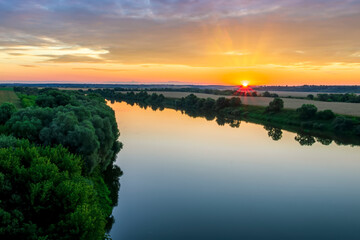 Naklejka na ściany i meble Scenic view at beautiful sunset or sunrise on a shiny river with green bushes on sides, golden sun rays, calm water ,deep blue cloudy sky and forest on a background, spring landscape