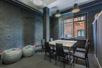 An atmospheric loft room with gray brick walls and a window, furnished with soft poufs, a table and chairs.