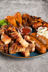 Assorted grilled meat in a plate on the table. Fried chicken legs and wings, sausages, shish kebab, pickled onions and tomato sauce. Vertical orientation, close-up, no people