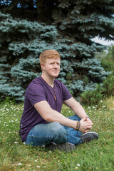 Naklejka na ściany i meble Portrait of redhead caucasian man wearing purple t-shirt and blue jeans and sitting on the grass in city park during lunch break at summer. Person looks away. Theme of rest in work break.