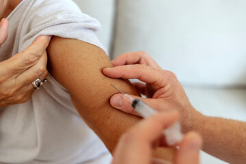 An male doctor is administering the COVID-19 vaccine to a senior woman. They are both wearing a face mask at the doctor's office. The doctor is slowly injecting the needle to the woman's arm.