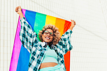Black woman with eyeglasses, headsets and afro hair holds up the lgbtq flag of gay pride that moves with the wind on a sunny day. She fights for sexual freedom. - obrazy, fototapety, plakaty