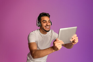 Handsome young Arab man wearing headphones, looking at tablet screen, using modern gadget in neon light