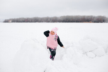 Fototapeta na wymiar kid who throwing snowball at camera, smiling and looking into camera wearing pink winter clothes in park. Astonishing background full of white color and snow. 