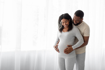 Fototapeta premium Satisfied millennial black handsome hugs pregnant wife in bedroom interior and look at belly