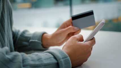 Close-up of female hands holding mobile phone and bank card shopping online. Unrecognizable woman pays for goods in web with smartphone enters secret account number book tickets makes order at table