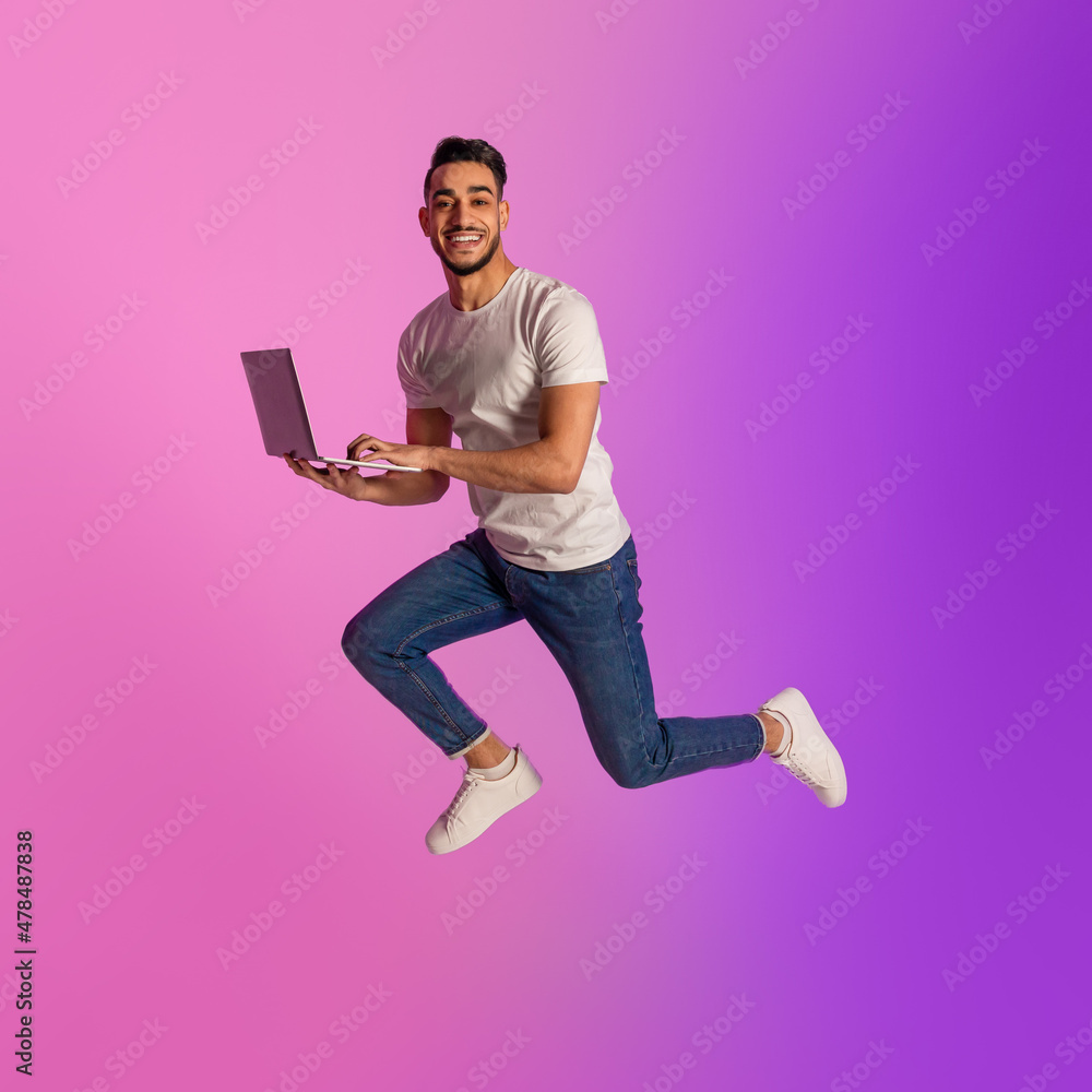 Poster full length of millennial arab man with laptop computer jumping up in neon light. cool new website o
