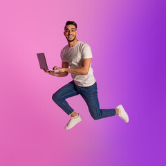 Full length of millennial Arab man with laptop computer jumping up in neon light. Cool new website or advertisement
