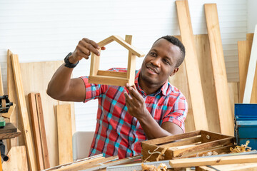 African black male carpenter man holding and checking wooden house model toy in carpentry workshop....